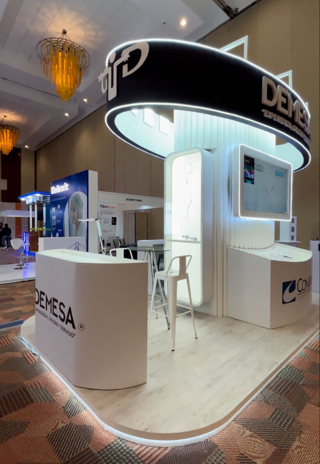 Stand custom con aro iluminado en el techo, panel retroiluminado, pantalla y mostradores sobre piso de madera.